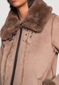 Brun faux suede-jacka med en mjuk, brun pälskrage, utrustad med en svart dragkedja och dekorativa tryckknappar framtill.