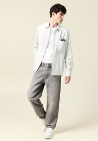 Jasnoszara koszula z guzikami i kieszonką, zestawiona z luźnymi szarymi jeansami i białymi sneakersami na neutralnym tle.