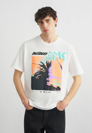 Νέος άνδρας με σγουρά μαλλιά που φοράει λευκό T-shirt της Nike με πολύχρωμο γραφικό φοίνικα, στέκεται με τα χέρια στις τσέπες.