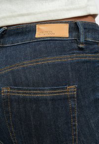 Rückansicht dunkler blaue Denim-Jeans mit oranger Naht und einem beigen Lederpatch mit der Aufschrift „Vero Moda We Do Denim“, getragen zu einem weißen Oberteil.