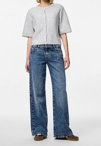 Světle šedý pletený kardigan s krátkým rukávem, kulatým výstřihem a zapínáním na knoflíky, spárovaný s volnými, bledě modrými džíny.