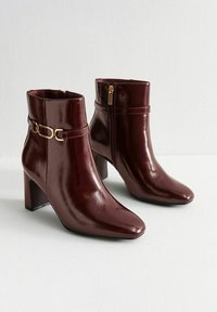 Botas de tobillo burdeos de charol con punta afilada, tacón cuadrado, cremallera lateral y detalle en tono dorado en la parte del tobillo.