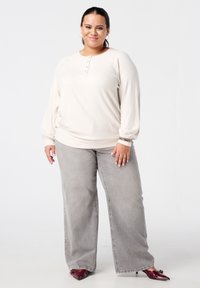 Glunderende vrouw in een crèmekleurige, knoopsluitende trui, grijze wijde jeans, hoepelletjes, armbanden en bordeauxrode puntige schoenen, staand op een witte achtergrond.