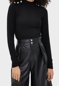 Kobieta w dopasowanej czarnej bluzce z długim rękawem i ribbowanym wzorem, z srebrnymi guzikami na ramieniu, połączonej z czarnymi skórzanymi spodniami o wysokim stanie z podwójnymi guzikami.