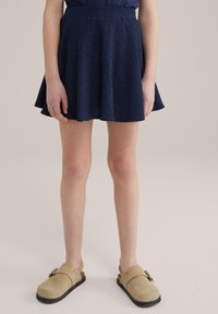 Marinblå, texturerad kjol med en utsvängd nederkant; bärs med ljusbruna träskor som har spänne. Bakgrunden är neutral och jämn.