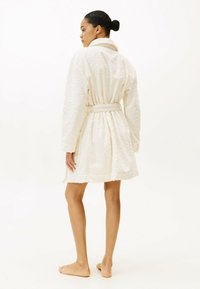 Accappatoio bianco con motivo paisley, maniche lunghe e vita annodata, che arriva a metà coscia. Modello mostrato di spalle, a piedi nudi.