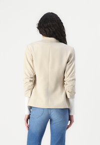 Blazer bej, croit cu mâneci căptușite, spate ajustat și design ușor structurat. Purtați peste o cămașă albă și asociați cu blugi albaștri deschiși.