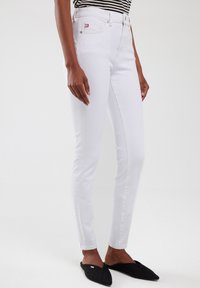 Jeans blancos ajustados de mezclilla, con cintura alta, presillas para cinturón y una pequeña etiqueta de logo en rojo. Combinados con zapatos negros tipo mocasín.