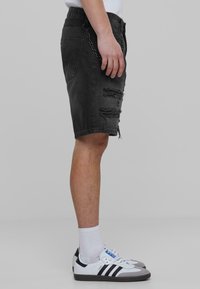 Shorts de mezclilla negros con bordes deshilachados y rotos en la parte frontal, combinados con zapatillas blancas con rayas negras y detalles grises.