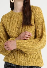 Sinappikeltainen neulepaita, jossa on teksturoitu kuvio, pitkät hihat ja pyöreä pääntie. Rentoon istuvuuteen ja resoroituun helmaan suunniteltu.