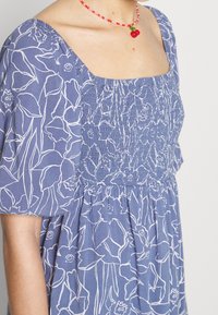 Femme portant une robe bleue avec des contours floraux blancs et des manches bouffantes, associée à un collier rouge en perles orné d'un pendentif cerise.