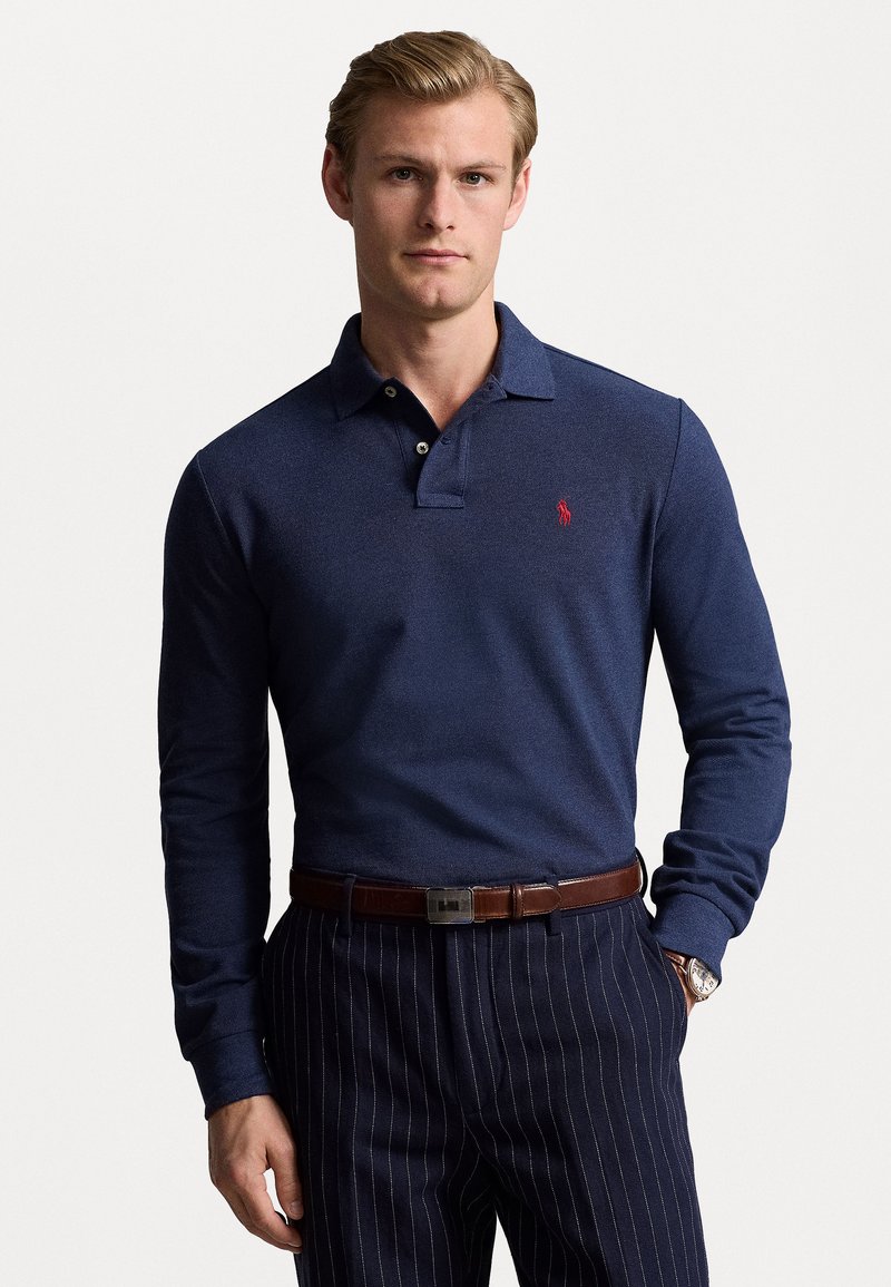 Marineblauw polo met lange mouwen en een gestructureerde afwerking, voorzien van een rode logo. Gecombineerd met gestreepte broek en een bruine riem. Eenvoudige knoopsluiting.