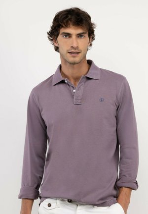 Hombre con pelo rizado y castaño, vestido con una camiseta polo morada de manga larga y pantalones blancos, de pie frente a un fondo liso y claro.