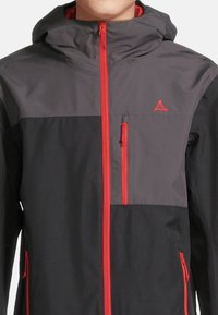 Schwarze und graue Kapuzenjacke mit roten Akzenten, ausgestattet mit einem Reißverschluss vorne, Seitentaschen und einem Logo auf der Brust. Das Material scheint leicht und glatt zu sein.