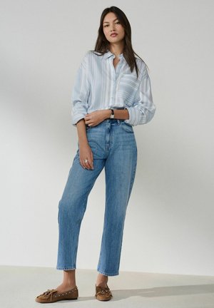 Junge Frau steht und trägt ein hellblaues, vertikal gestreiftes Hemd, hoch taillierte blaue Jeans, braune Loafers und eine Armbanduhr am linken Arm.