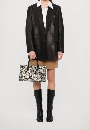 Femme portant un blazer en cuir noir à double boutonnage, une chemise blanche, une jupe beige, des bottes noires montant jusqu'au genou, tenant un sac à main noir et blanc à pois.