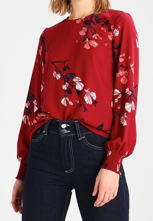 Femme portant un chemisier rouge à fleurs à manches longues, rentré dans un jean bleu foncé taille haute avec des doubles boutons à l'avant.