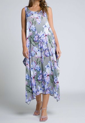 Femme portant une robe grise sans manches avec de grands motifs floraux bleus et violets, assortie à des sandales à lanières violet clair.