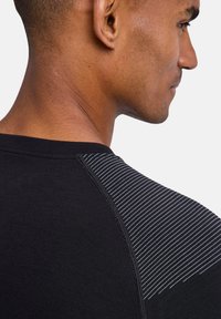 T-shirt de sport noir avec un tissu texturé à finition lisse et un accent rayé sur l'épaule. Vue rapprochée de côté.