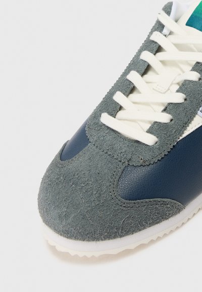 Sneaker cu un superior din suede gri, accente din piele bleumarin, sireturi albe și o talpă din cauciuc texturat. Vârf rotunjit și detalii cusute.