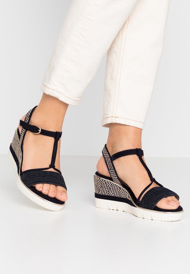 Pieds chaussés de sandales compensées noires tissées avec brides à la cheville et semelle blanche, assorties à un pantalon court écru.
