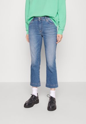 Persoon draagt cropped blauwe spijkerbroek, witte sokken, zwarte veterschoenen en een groene trui tegen een effen lichte achtergrond.