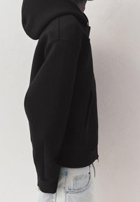 Personne portant un sweat à capuche noir zippé avec la capuche relevée, associé à un jean en denim bleu clair, debout contre un mur clair uni.