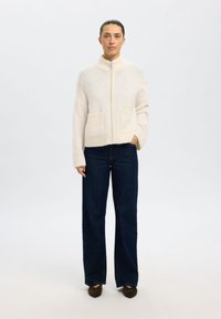 Suéter crema con cremallera y cuello alto, textura suave y dos bolsillos delanteros, combinado con jeans oscuros de pierna ancha y zapatos oscuros.