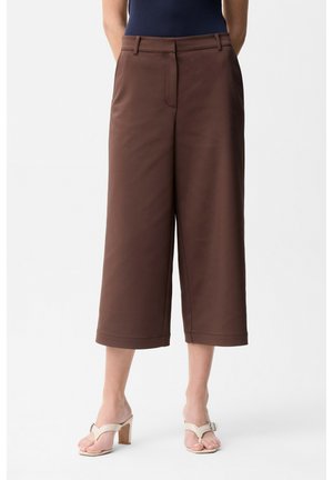 Femme portant un pantalon marron taille haute, coupe large et court avec fermeture devant, associé à des sandales à talons beiges et un haut sans manches foncé.