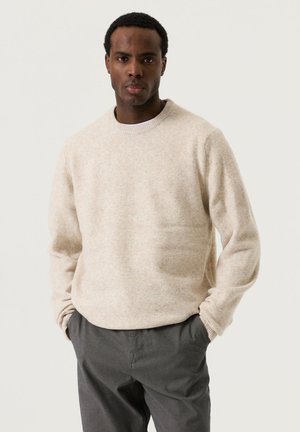 Man wearing a light beige crewneck sweater and dark gray pants stands with hands in pockets against a plain white background.
