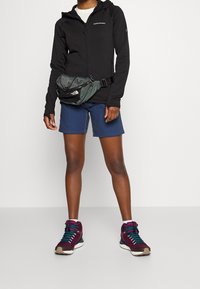 Chaqueta negra con cremallera, pantalones cortos azules y zapatillas altas burdeos con cordones turquesa. Se lleva una riñonera gris en la cintura.