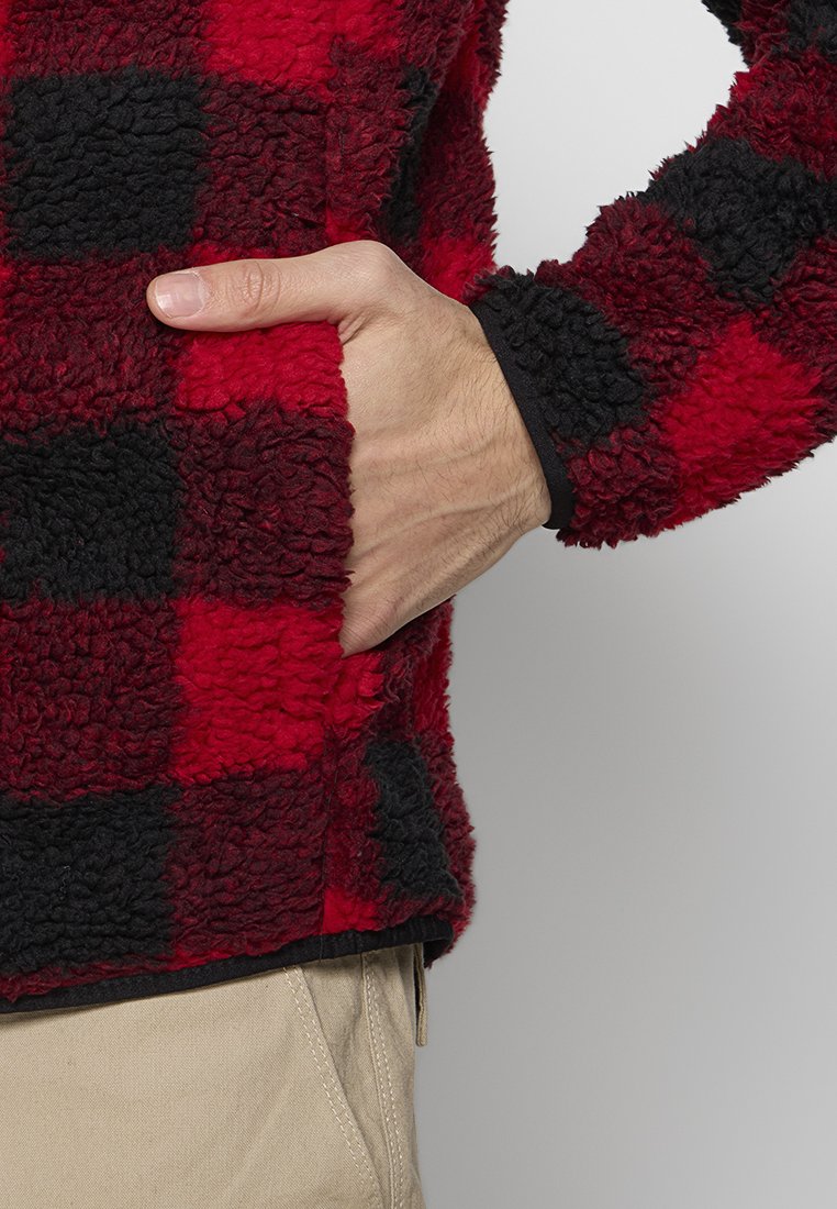 Czerwono-czarny, w kratkę, polarowy kurtka z miękką, teksturowaną powierzchnią; ma boczną kieszeń oraz czarne wykończenia przy mankietach i dolnej krawędzi.
