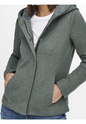 Vrouw draagt een groen capuchonjack met rits en drukknopen, handen in de zakken, over een wit overhemd en blauwe spijkerbroek.