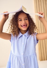 Trumpomis rankovėmis mėlynos ir baltos langeliais margintos suknelės su sagomis priekyje dizainas, kurią virš galvos laiko garbanotas vaikas ir šypsosi.