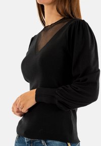 Maglione nero a coste con scollatura profonda a V e yoke in rete trasparente. Le maniche a palloncino aggiungono texture; il corpo aderente esalta la silhouette.