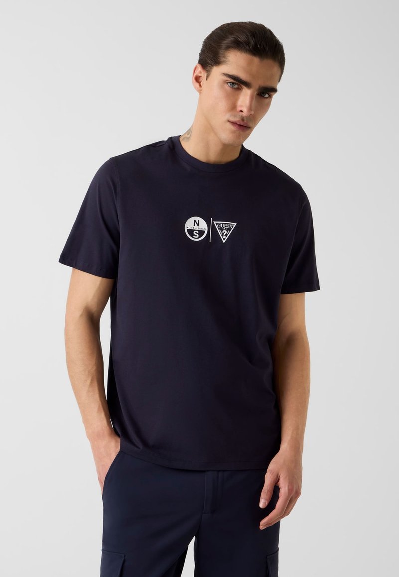 Homme portant un t-shirt bleu marine avec les logos North Sails et Guess sur la poitrine, debout les mains dans les poches devant un fond uni.