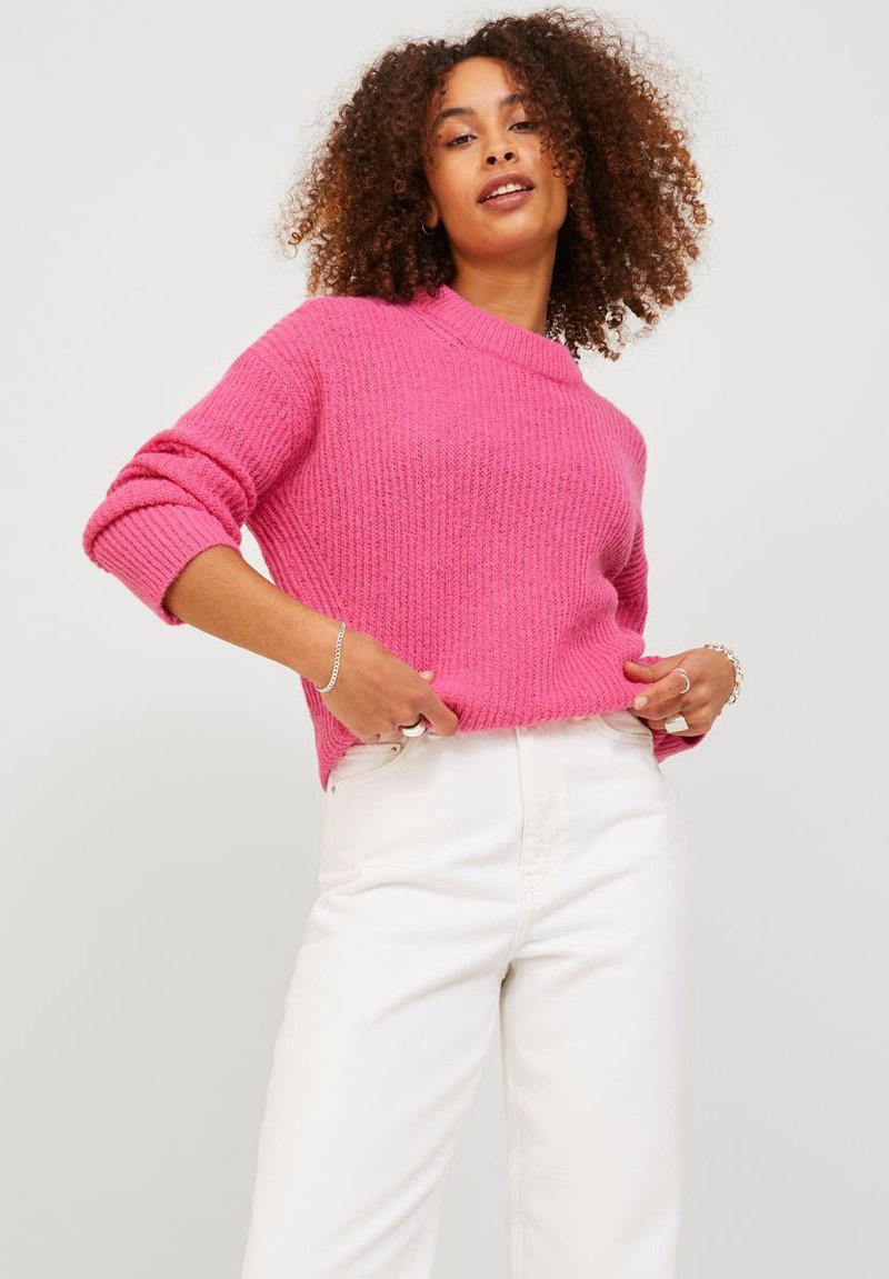 Pink knitted sweater with a textured pattern, high neckline, and rolled sleeves, paired with white trousers.