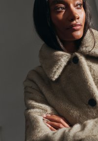 Beige jas van imitatiebont met een brede kraag, voorzien van zwarte knopen en een gestructureerd oppervlak. Model met gekruiste armen, waarbij de nagellak zichtbaar is.