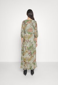 Langes, locker sitzendes Kleid mit floralem Muster in Grün, Pink und Beige. Hat Dreiviertelärmel und einen Rüschensaum. Wird mit schwarzen Knöchelstiefeln getragen.
