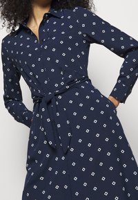 Nainen, jolla on päällä tummansininen pitkähihaisen mekko, jossa on valkoisia ruutukuviota ja vyötäisillä solmittu vyö, kädet taskuissa.