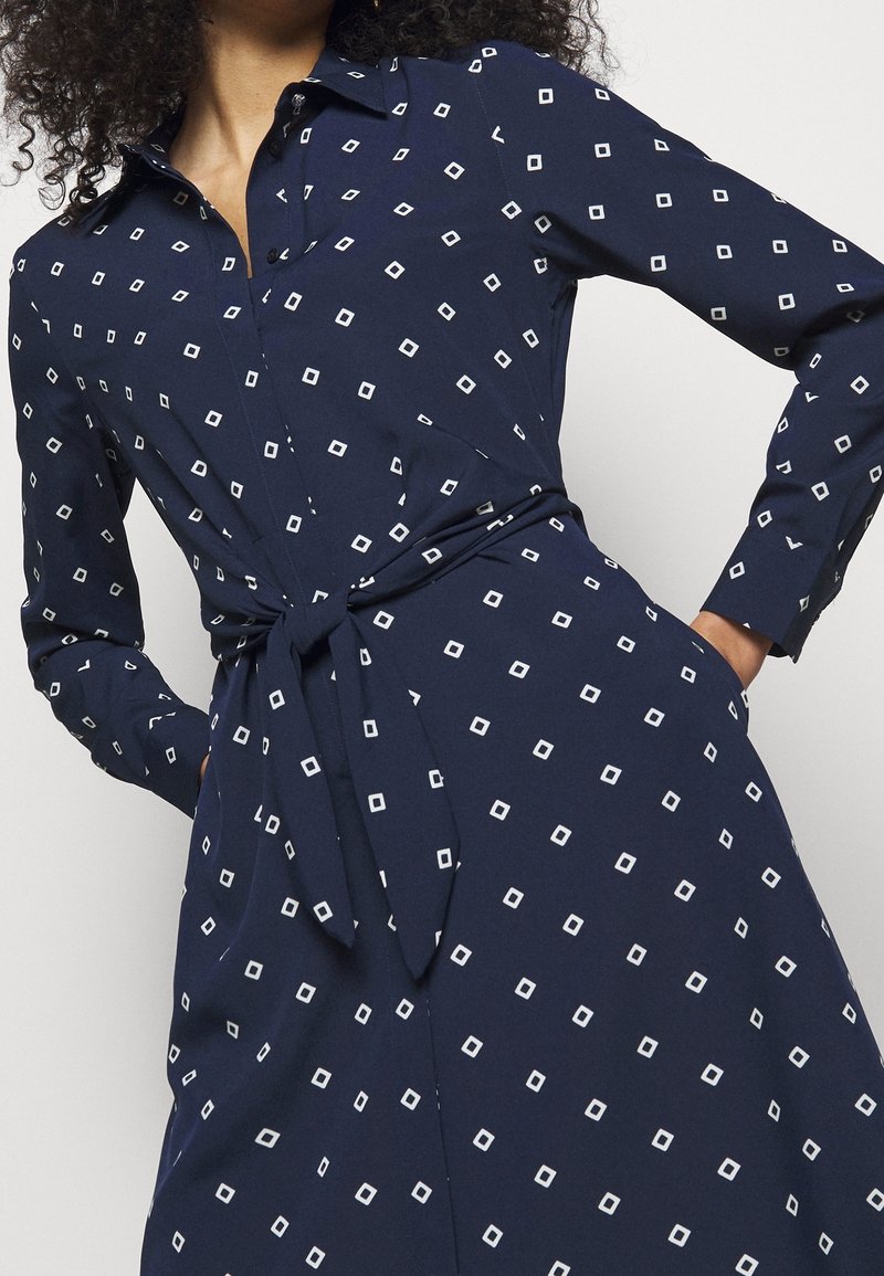 Nainen, jolla on päällä tummansininen pitkähihaisen mekko, jossa on valkoisia ruutukuviota ja vyötäisillä solmittu vyö, kädet taskuissa.