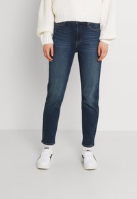 Dark blue denim jeans with a slim fit, five pockets, and minimal stitching. Paired with white knitted sweater and white sneakers.