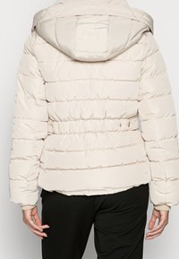 Person som bär en ljusbeige puffjacka med huva, står med ryggen mot kameran, i kombination med svarta byxor.