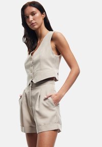 Gilet beige su misura con sei bottoni e tasche abbinato a pantaloni corti a vita alta con pieghe e risvolti.