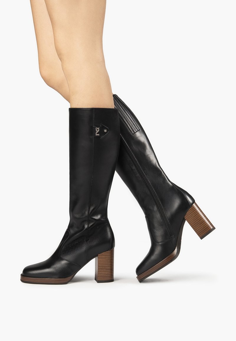 Jambes chaussées de bottes en cuir noires montant jusqu'aux genoux, avec talons blocs en bois empilés et fermeture éclair latérale, sur fond blanc.