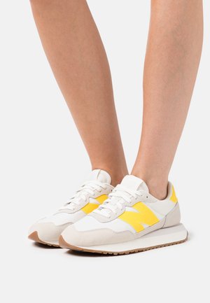 Białe i żółte sneakersy z cholewką z zamszu i siateczki, z charakterystycznym zakrzywionym kształtem, gumową podeszwą oraz wyraźnym logo "N" na boku.