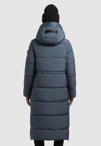 Lang, fyldig, mørkeblå frakke med hætte. Har vandrette quiltede sektioner og en sort logodetalje på ryggen. Sort strikhue båret.