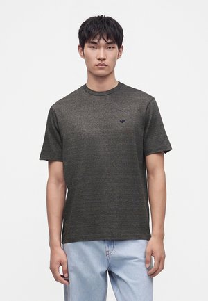 Jeune homme aux cheveux noirs et courts portant un t-shirt texturé à manches courtes gris foncé et un jean bleu clair, debout devant un fond uni.
