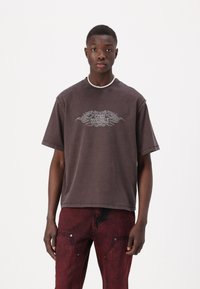 Brūns oversize T-krekls ar "Guess Originals" grafiku centrā, īsām piedurknēm un brīvu piegriezumu; valkāts ar sarkaniem nodilušiem džinsiem.