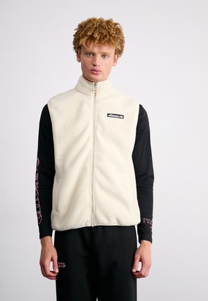 Cream-colored fleece vest with a high collar, front zipper, and logo patch, worn over a black long-sleeve shirt and black pants.